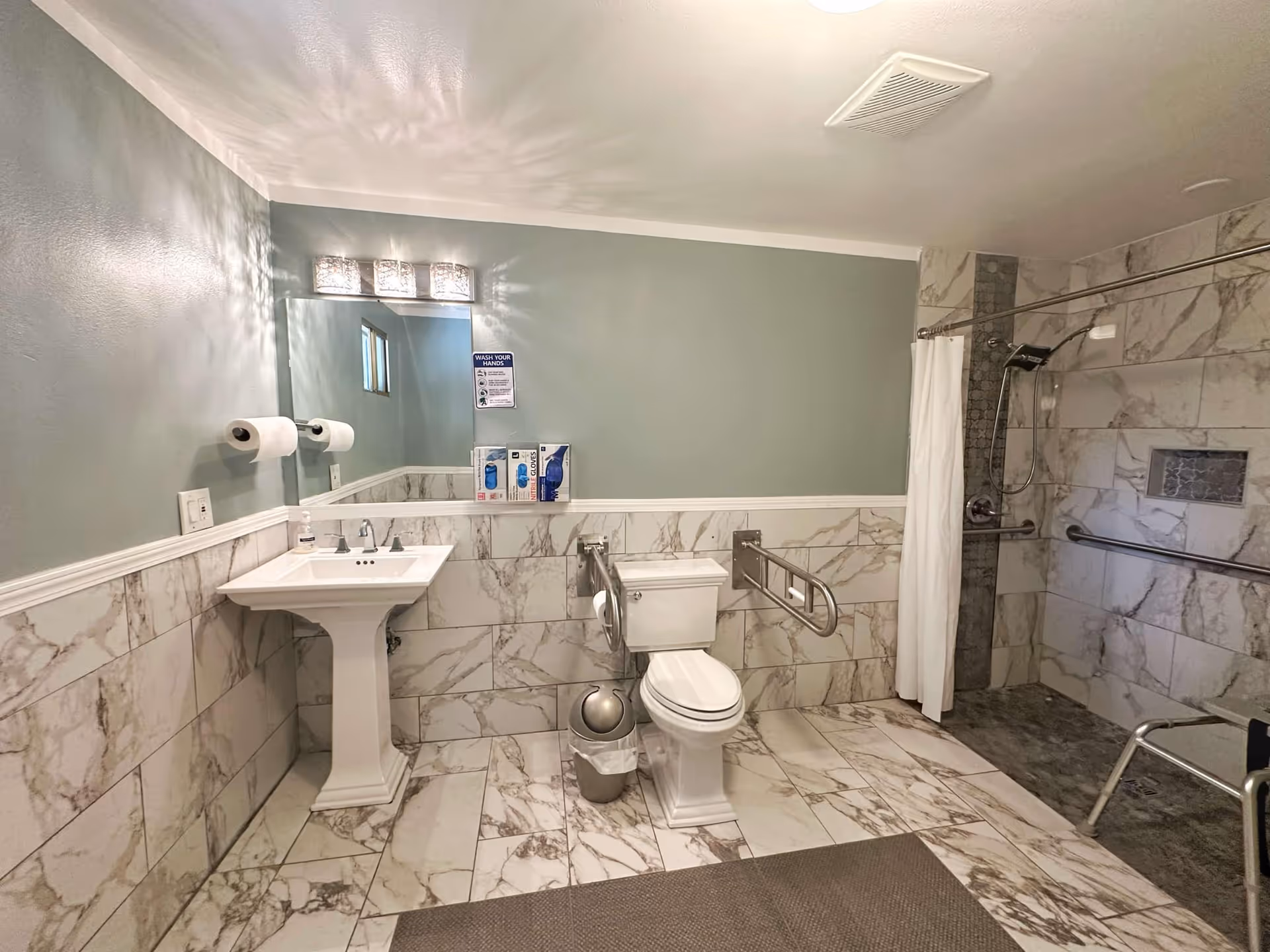 Accessible facility bathroom with pedestal sink, toilet with grab bars, mirror, and a tiled shower.