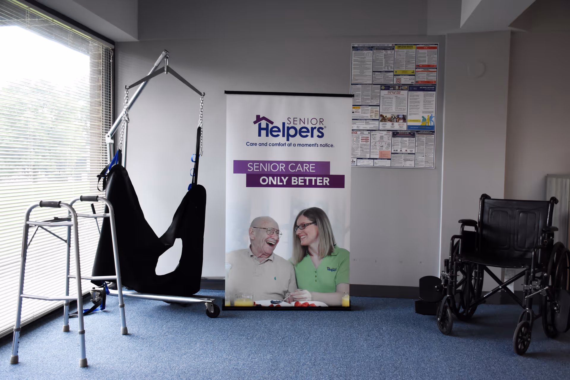 Indoor facility room with a patient lift, walker, wheelchair, and Senior Helpers banner.
