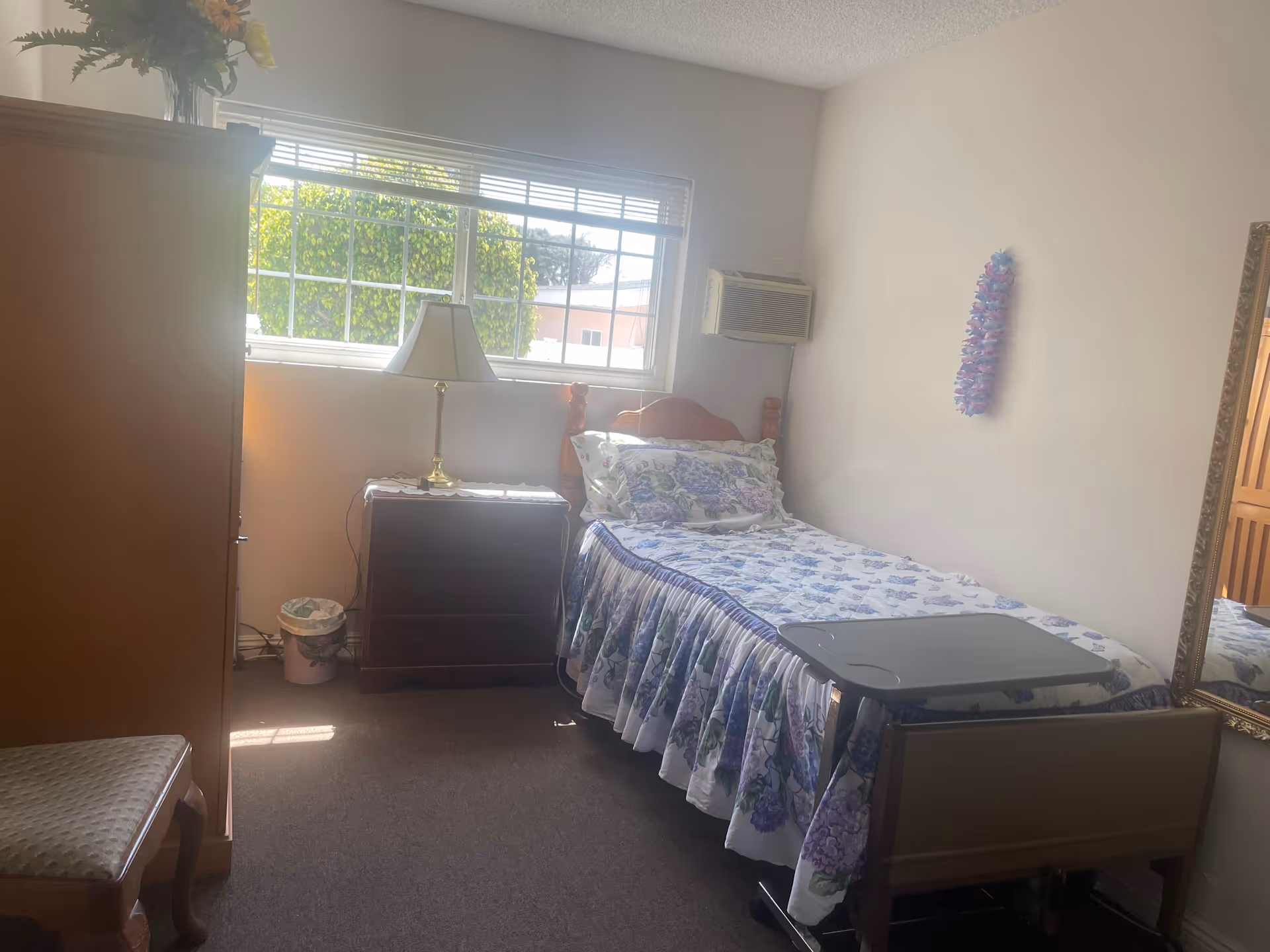 A small bedroom with a single bed, dresser, lamp, and window in a senior care facility.