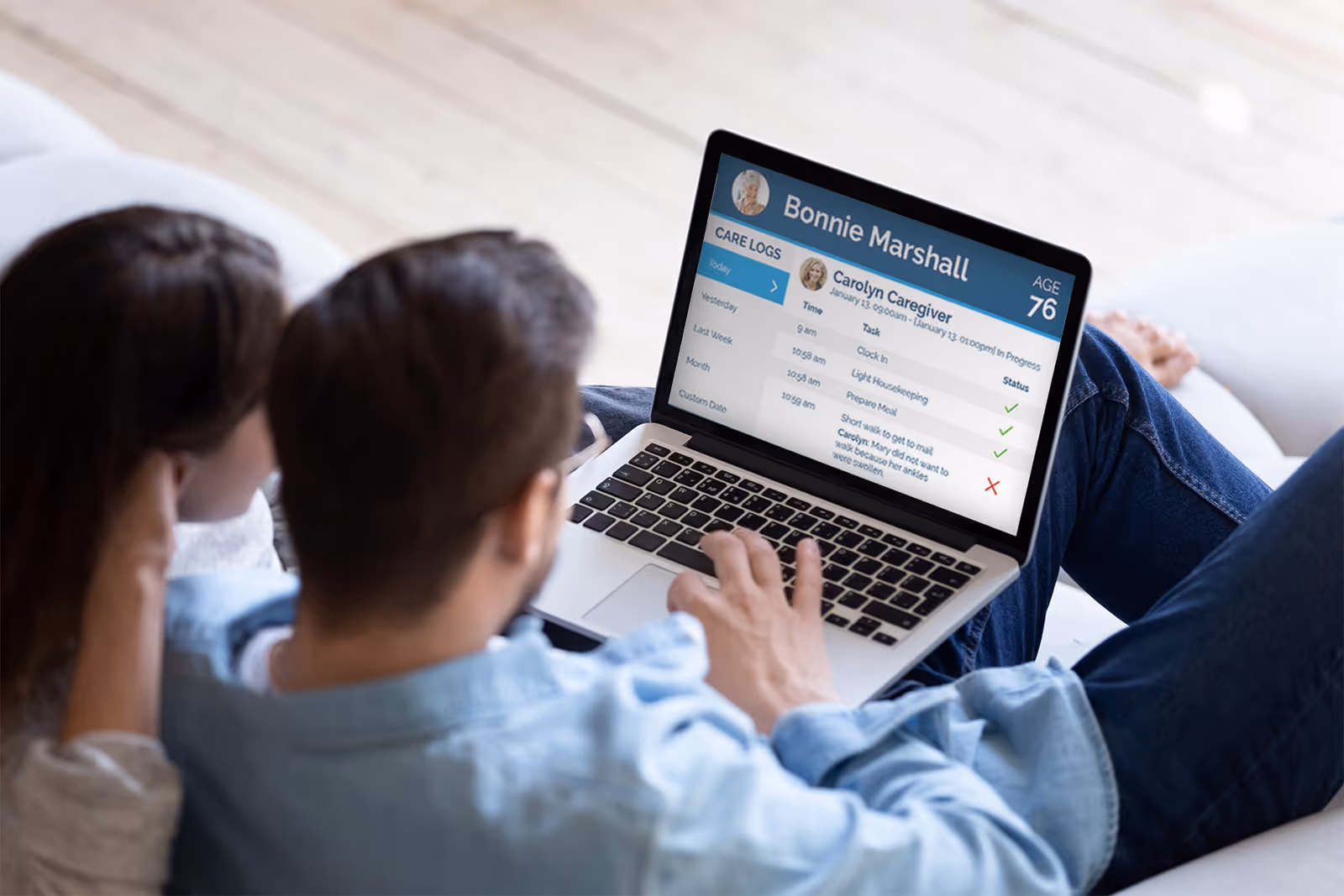 Couple sits on a living room sofa viewing a laptop displaying care logs.