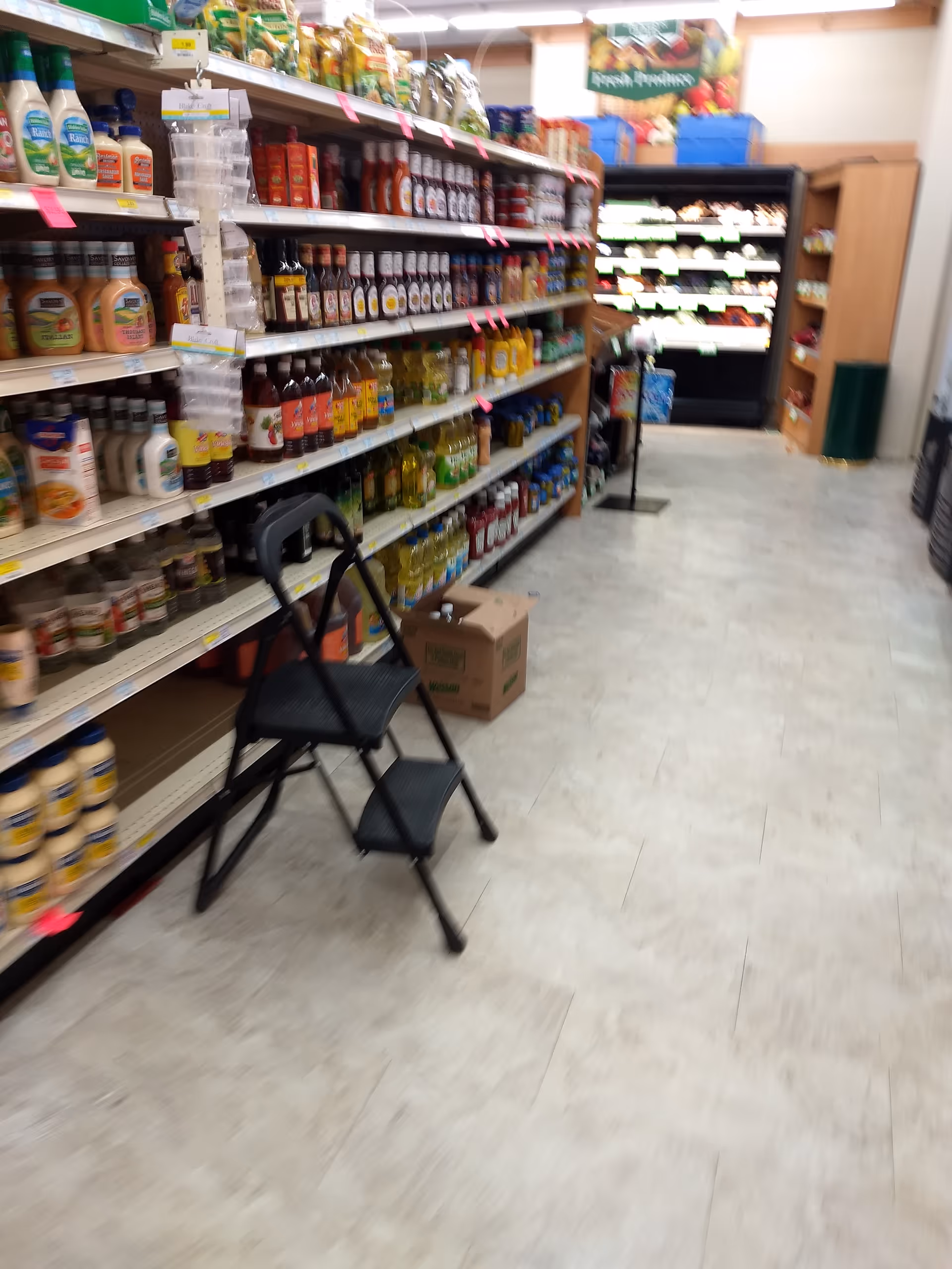 Long store aisle with shelves stocked with beverages and cleaning products, a step stool in foreground.