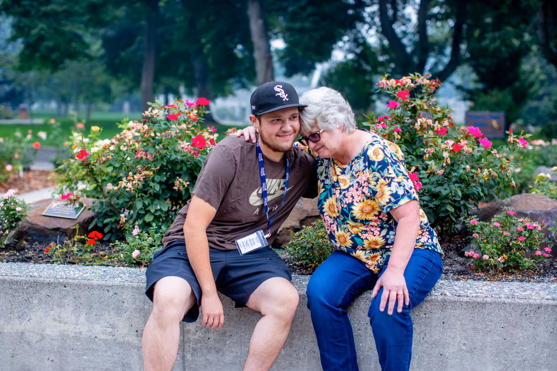 Two people sit on a concrete garden wall among blooming roses outside a senior care facility