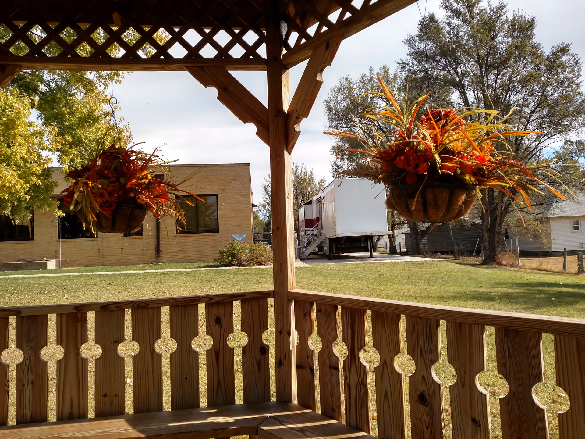 Wooden gazebo with hanging flower baskets over a grassy lawn and brick building.