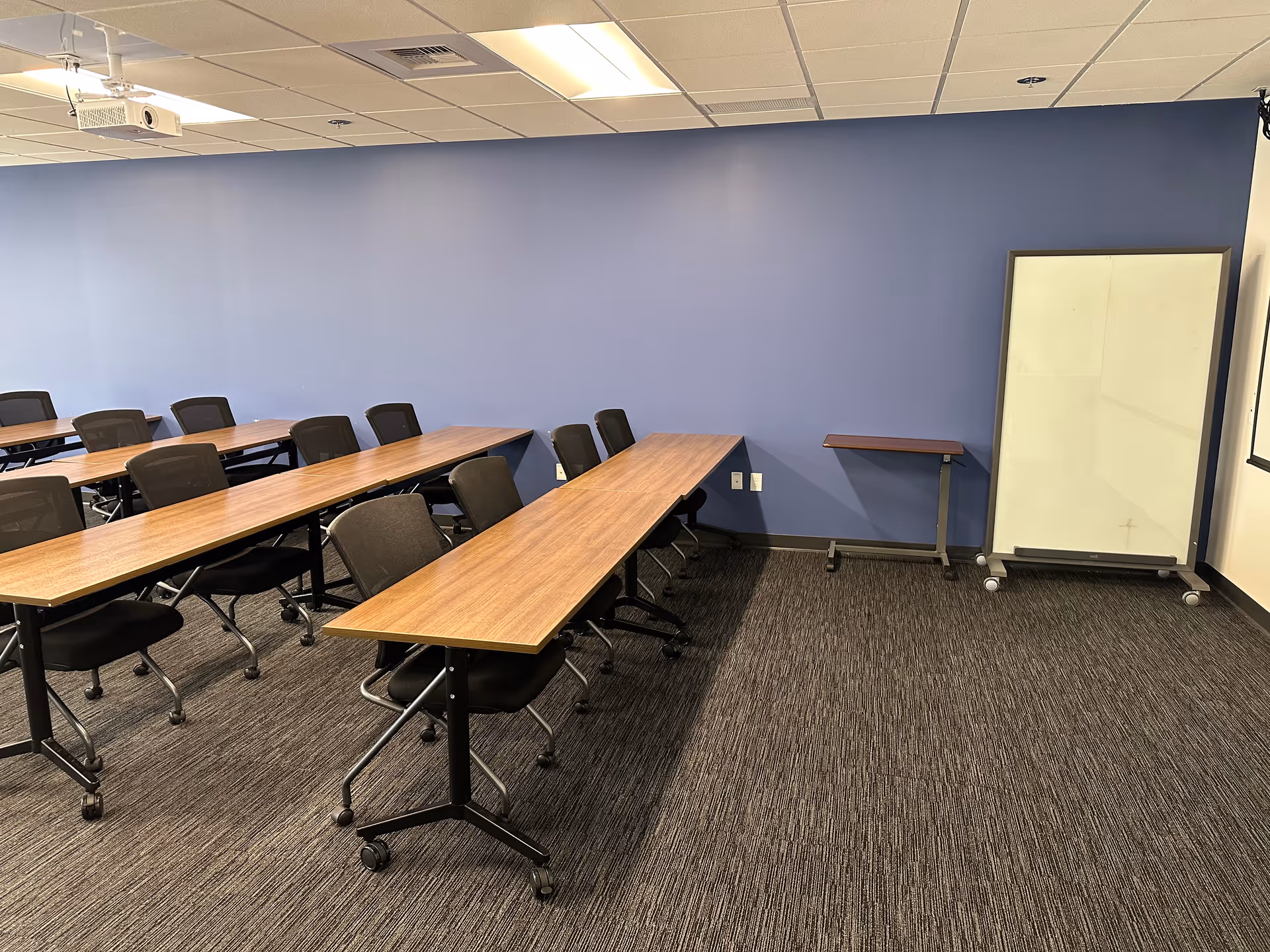 Blue-walled conference room with long wooden tables, black rolling chairs, and a mobile whiteboard.