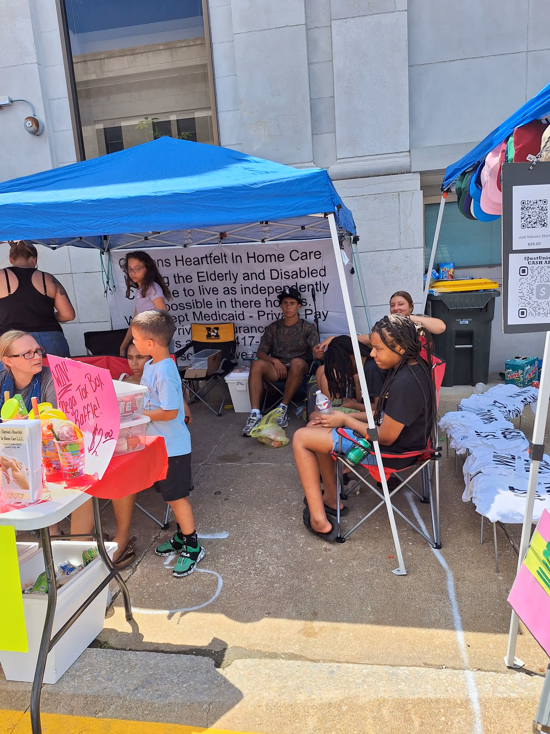 Outdoor charity booth with people under blue tents beside a building and tables.