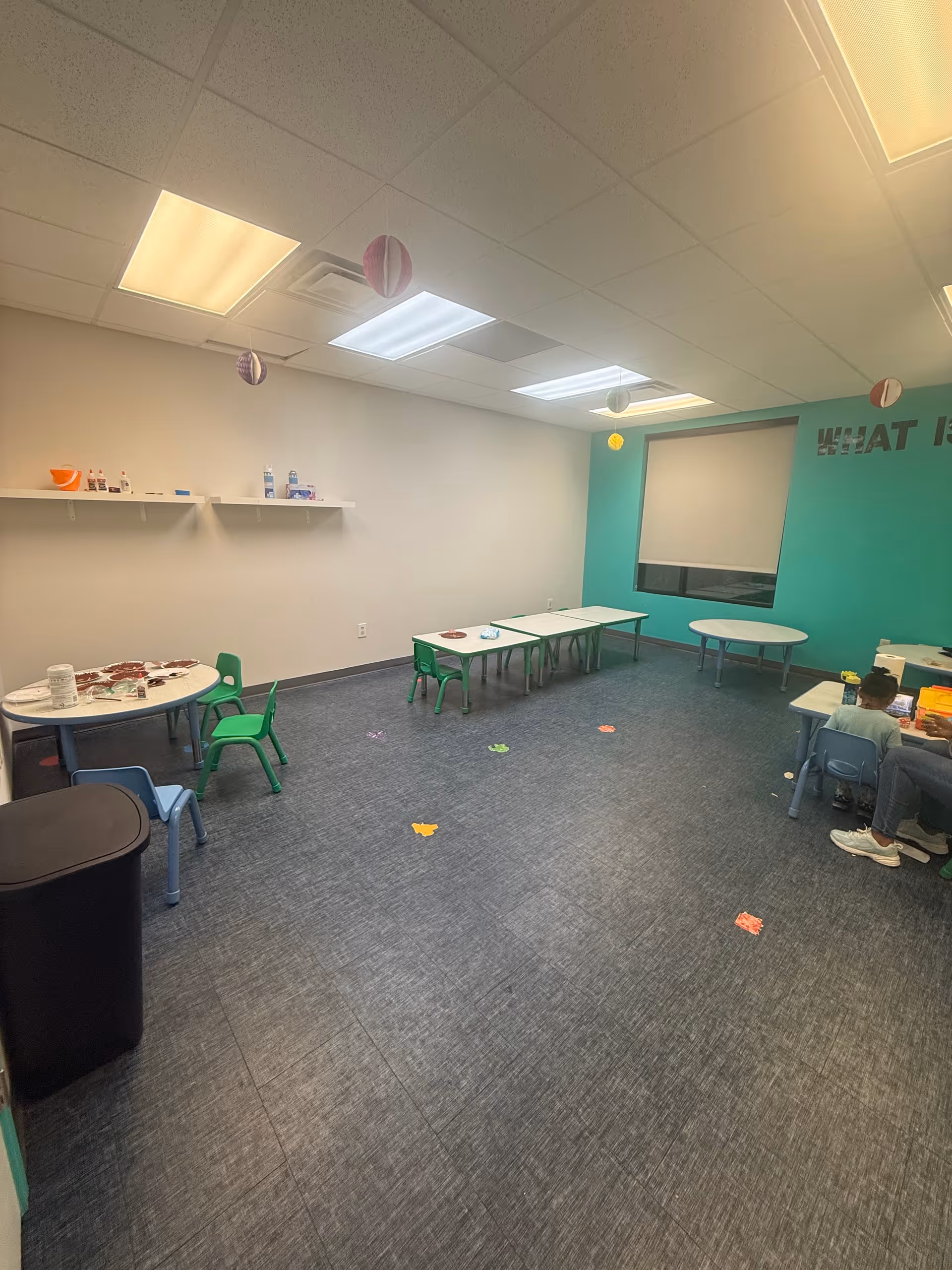 Bright therapy room with small tables, colorful chairs, teal wall, and hanging decorations.
