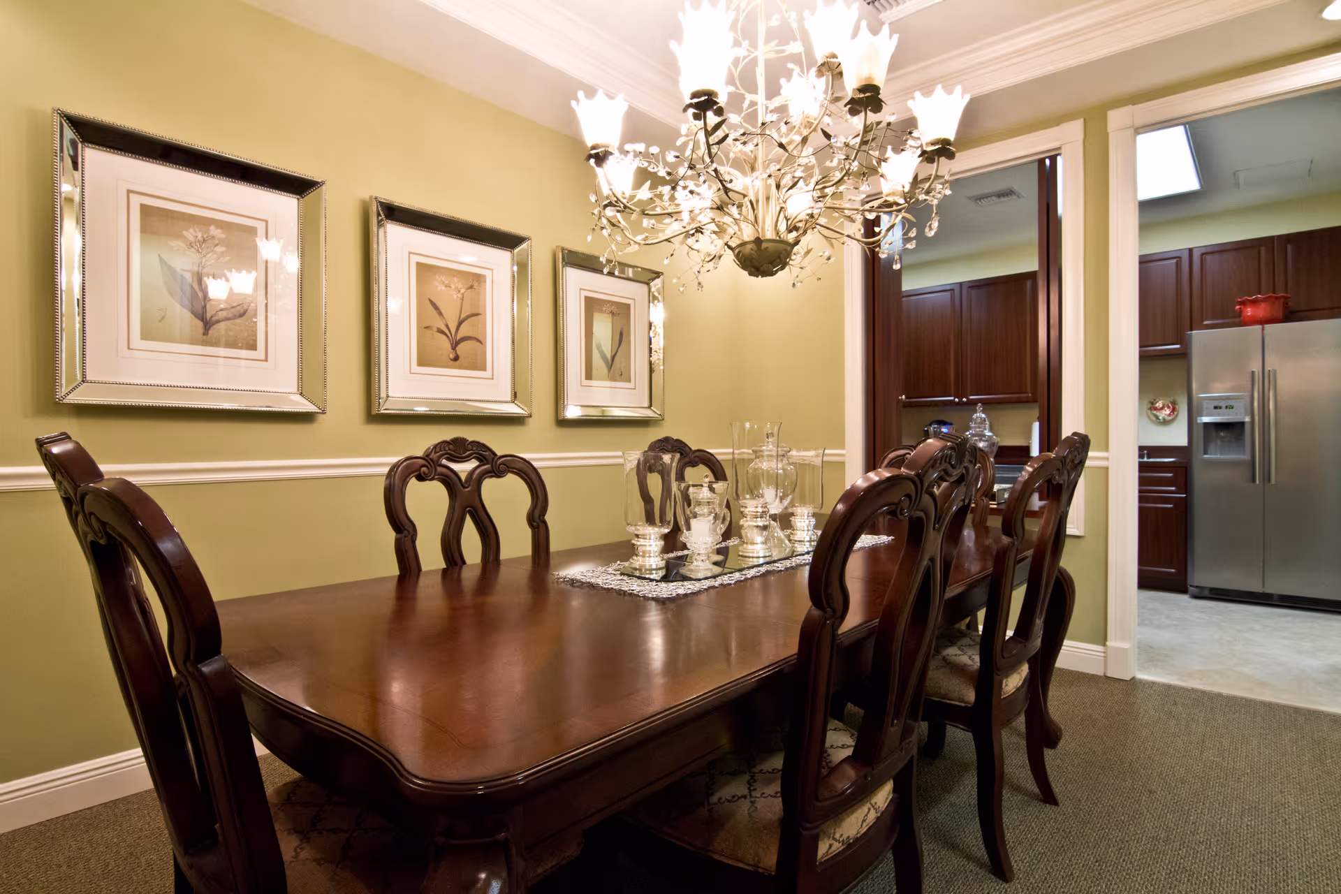 Formal dining room with dark wood table, chairs, chandelier, and kitchen visible in background.