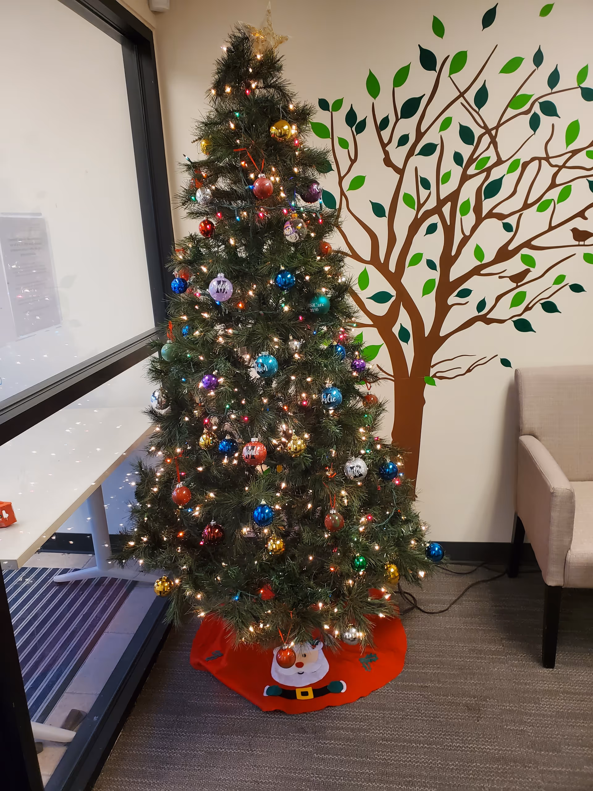 Decorated Christmas tree with colorful ornaments and lights stands beside a tree mural in a lounge.