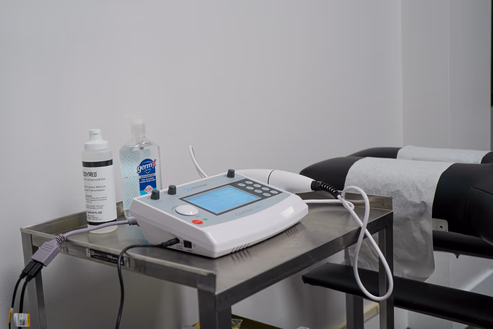 A medical device on a stainless steel cart beside a treatment bed in a clinical room.