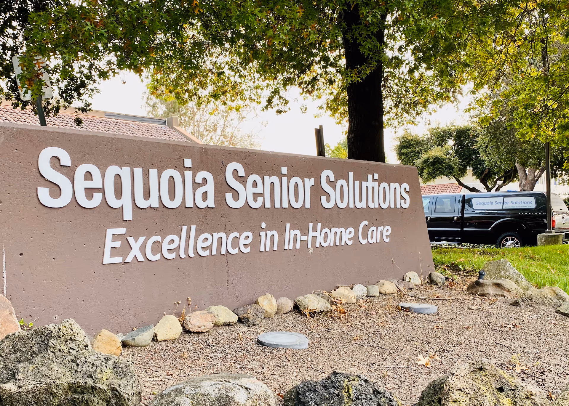 Exterior view of Sequoia Senior Solutions sign with trees and a black van nearby.