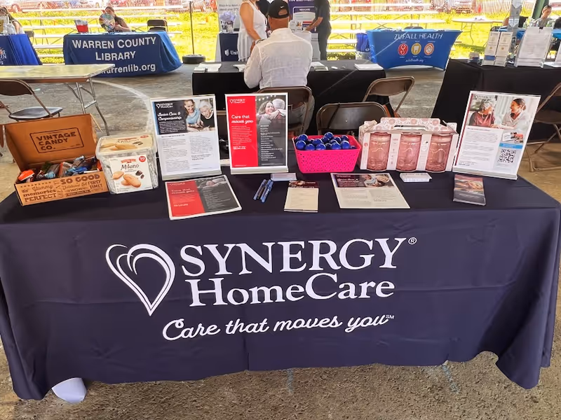 A Synergy HomeCare booth with a purple tablecloth and brochures at an outdoor event.