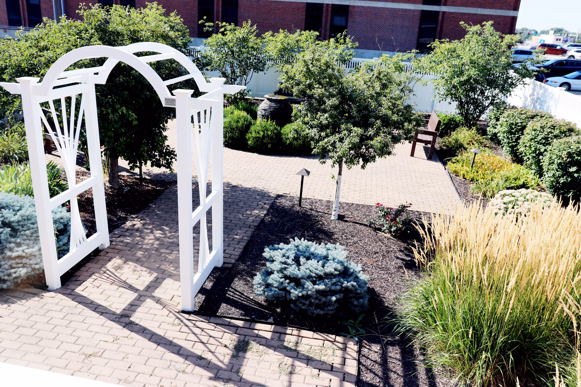 White garden arch over brick-pathed outdoor courtyard with trees and shrubs.