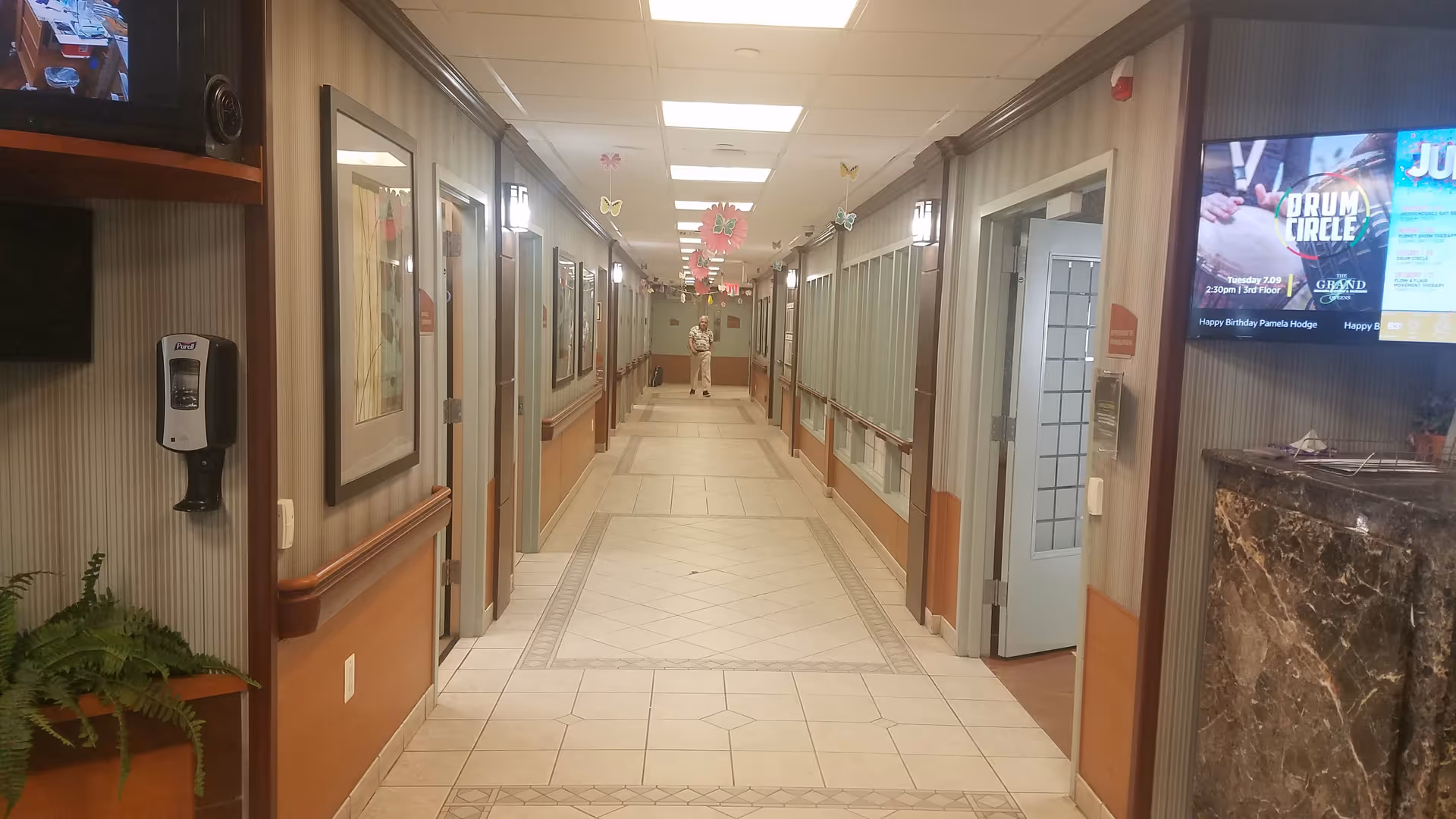 Long hallway in a senior living facility with doors, decor, and a person at the end.