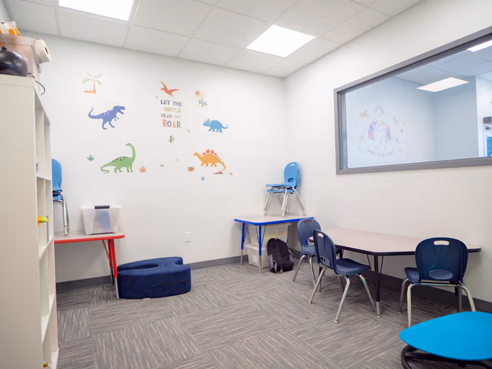 A bright therapy room with dinosaur wall decals, tables, and blue chairs.