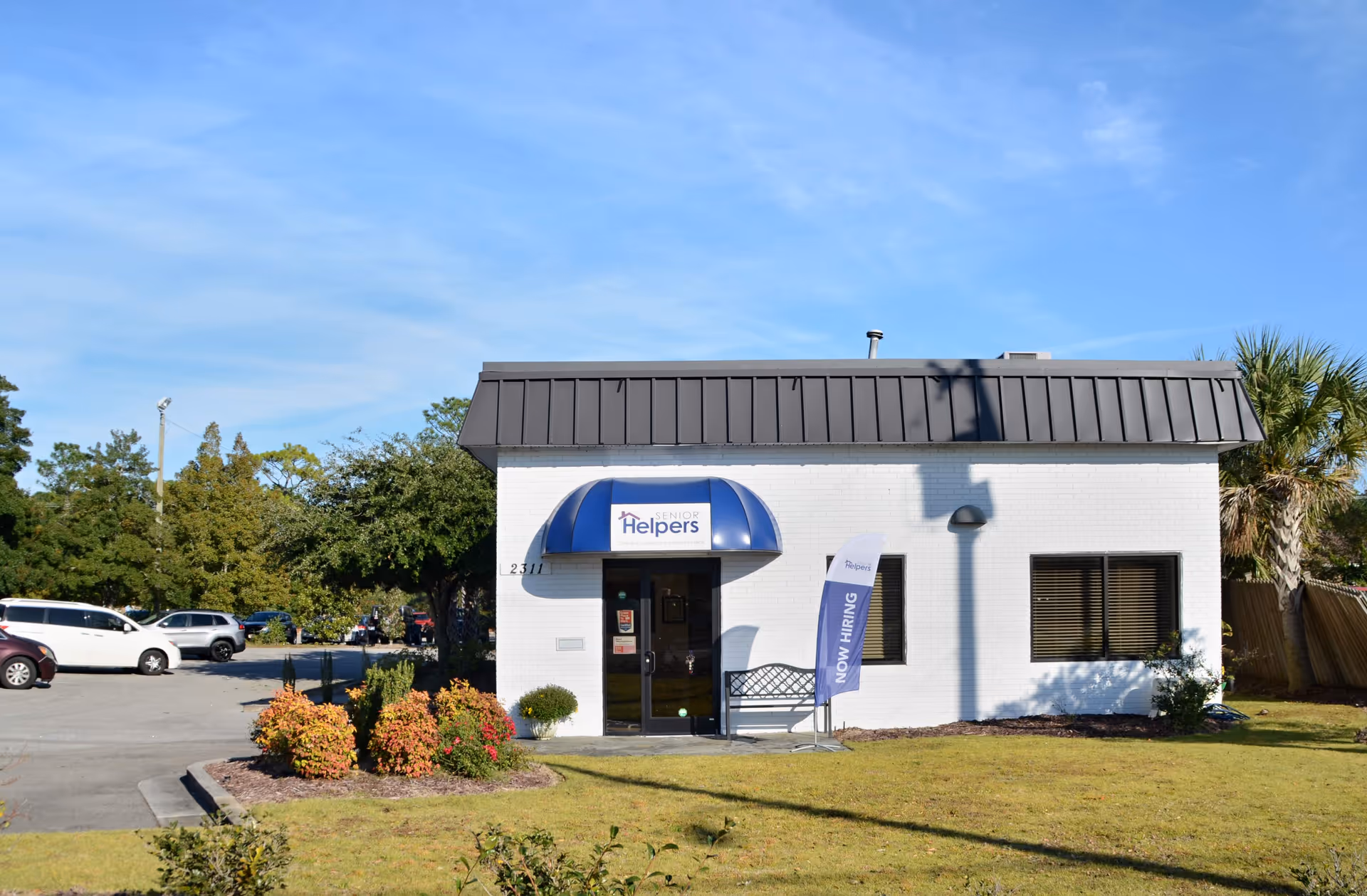 White single-story Senior Helpers facility with blue awning entrance, adjacent parking, and landscaping.