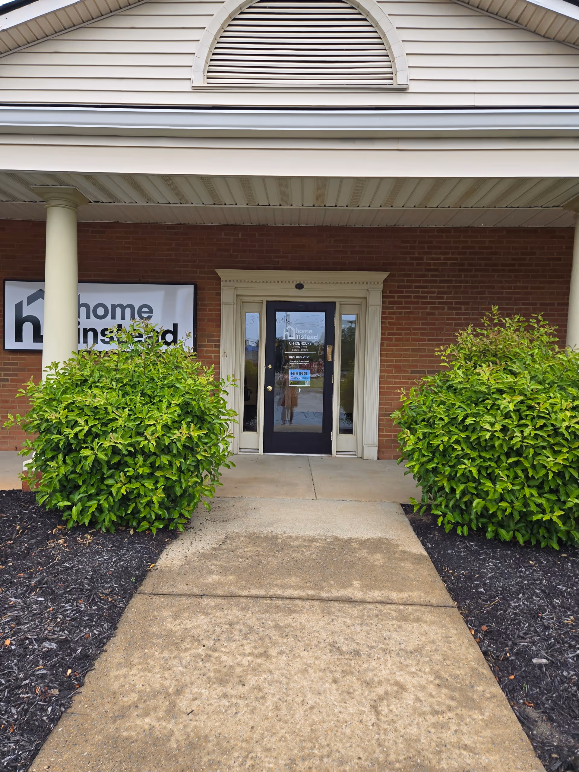 Entrance to a Home Instead facility with brick exterior, glass door, and green shrubs.