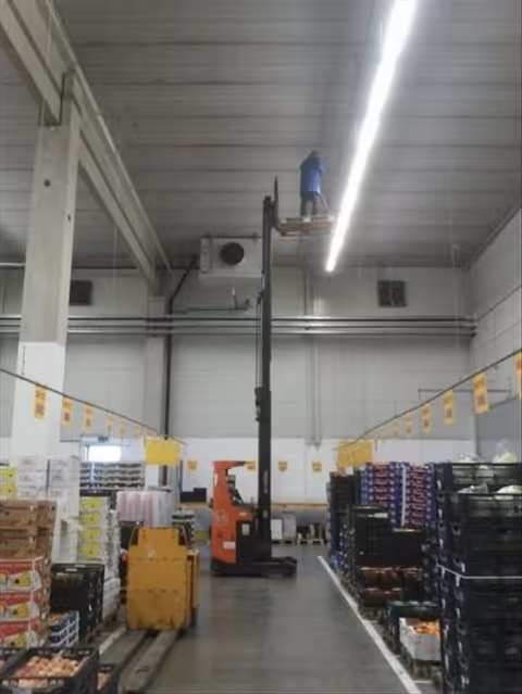 Industrial warehouse interior with tall ceiling, shelving, pallets, and a worker on a lift.