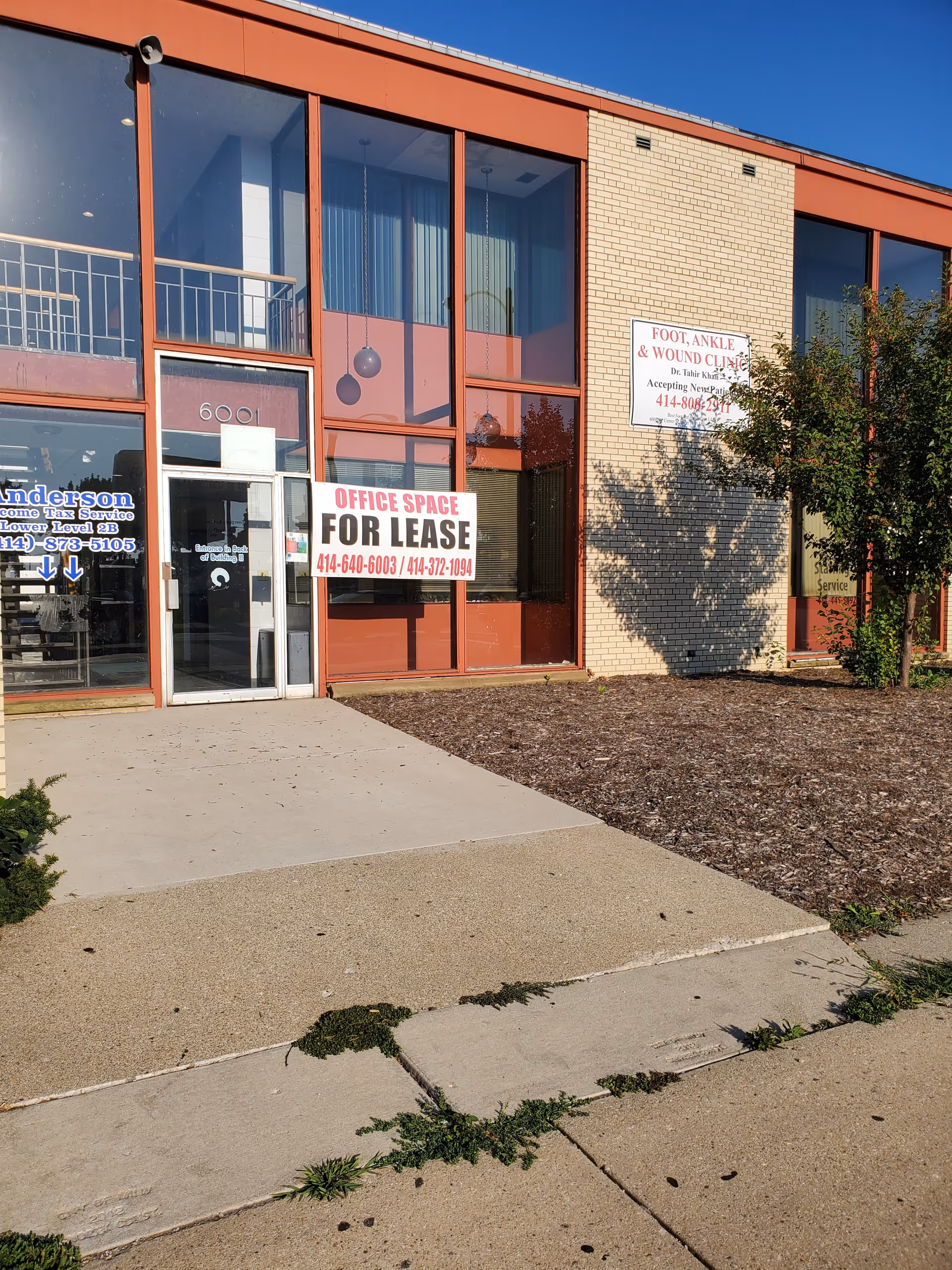 Exterior view of a brick and glass storefront with a large FOR LEASE sign and sidewalk.