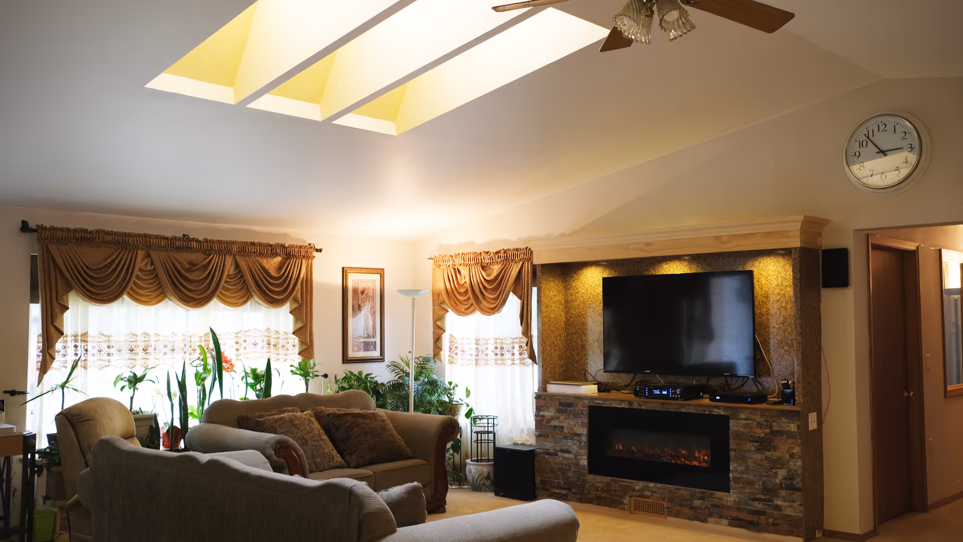 Bright living room with skylights, seating, plants, and a stone fireplace with a flat-screen TV.