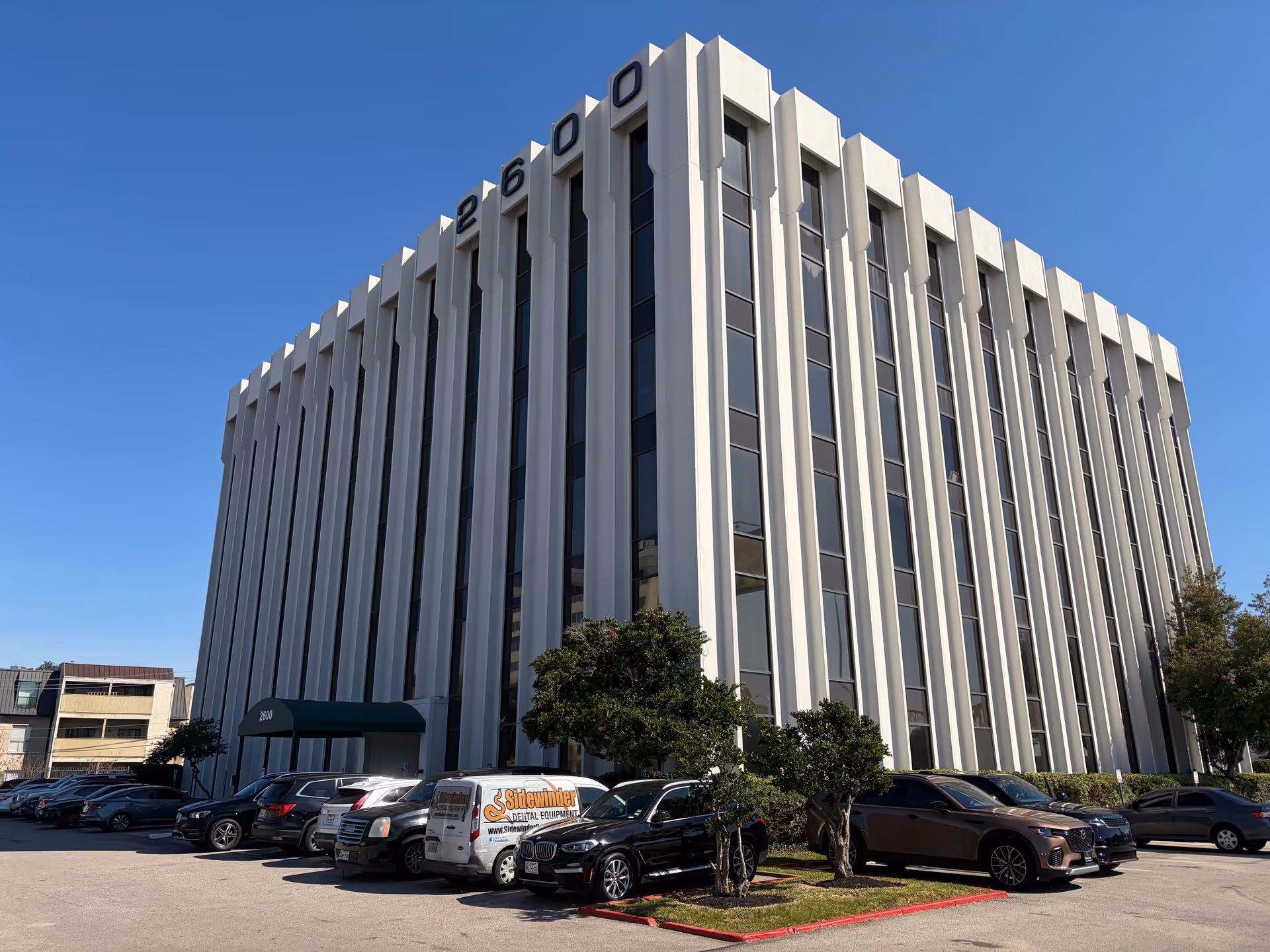 Tall white multi-story office building with vertical windows and a parking lot in front.