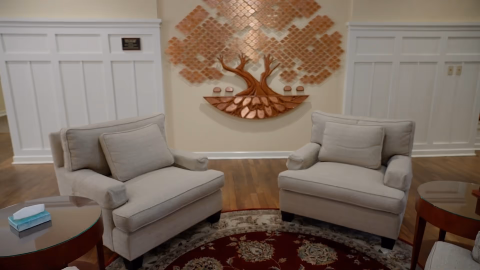 Two beige armchairs in a lobby with glass tables and decorative wall art.