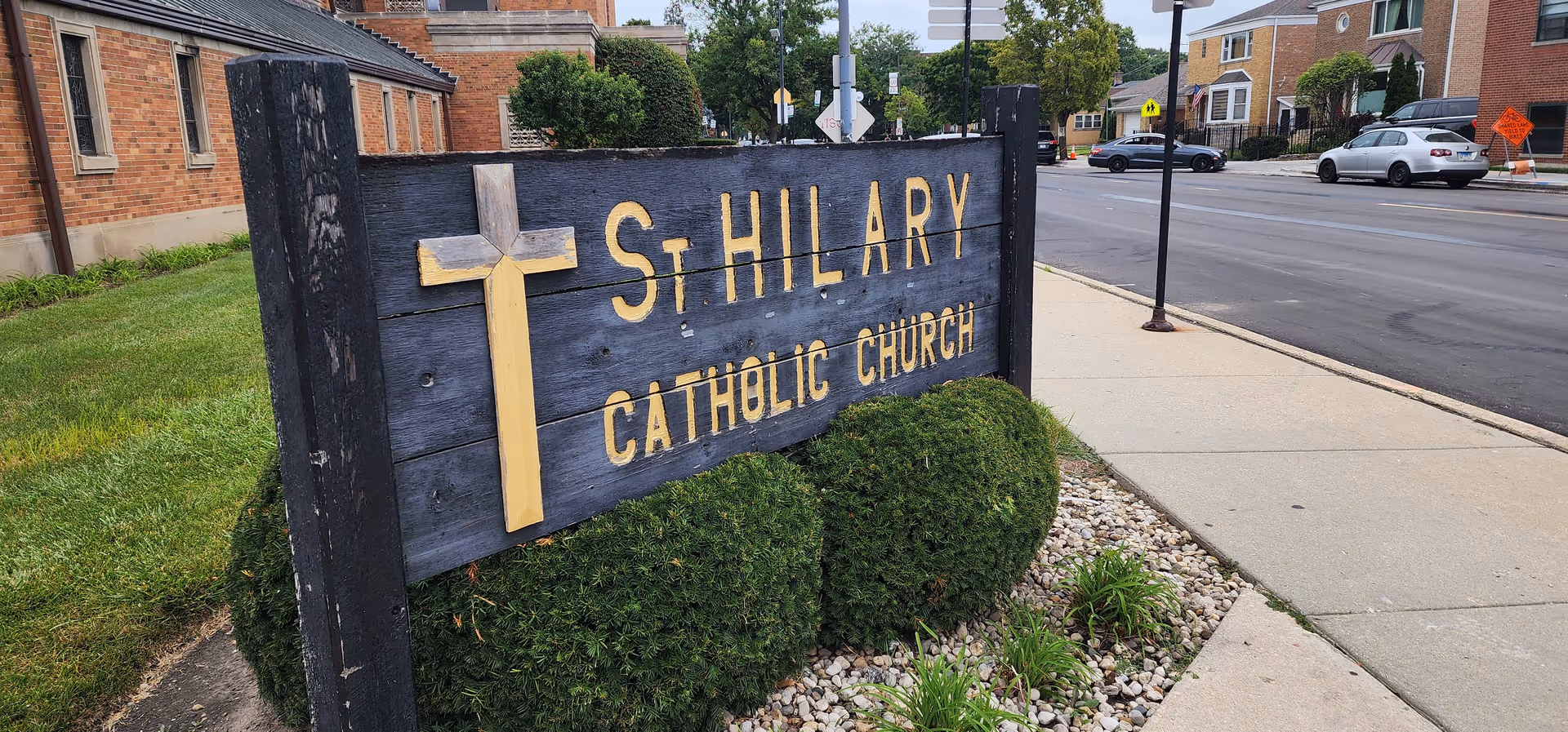 Wooden sign for St Hillary Catholic Church beside a sidewalk along a street