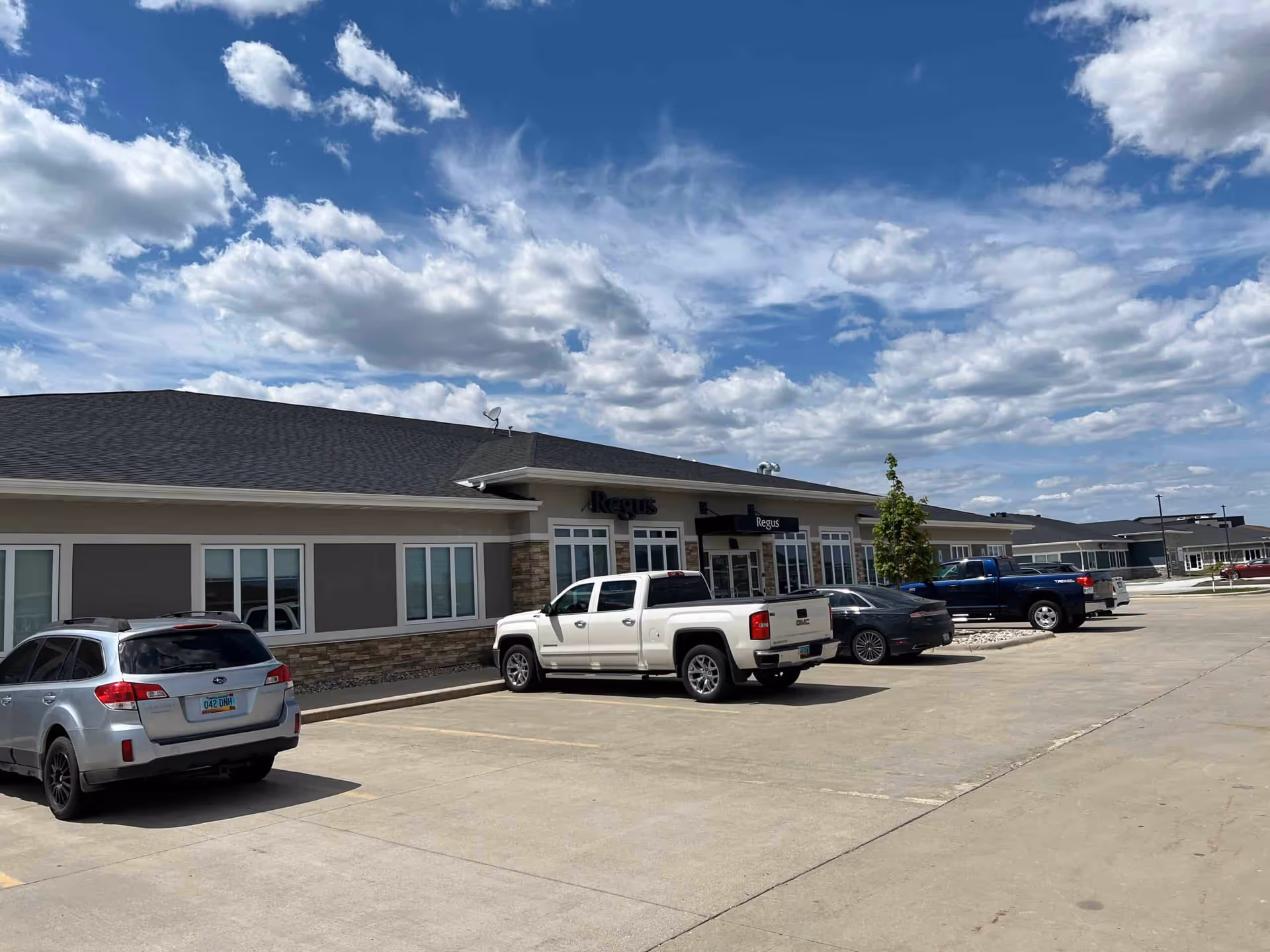 Exterior view of a single-story storefront building with parked cars and blue sky.