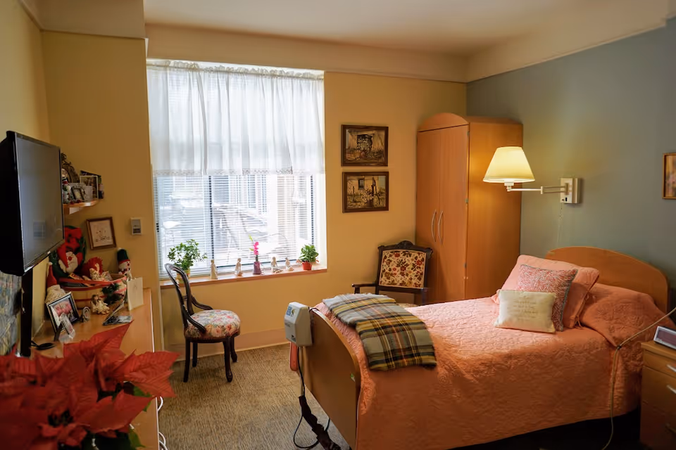 Cozy bedroom with pink quilt bed, window, wardrobe, lamp, and plants.