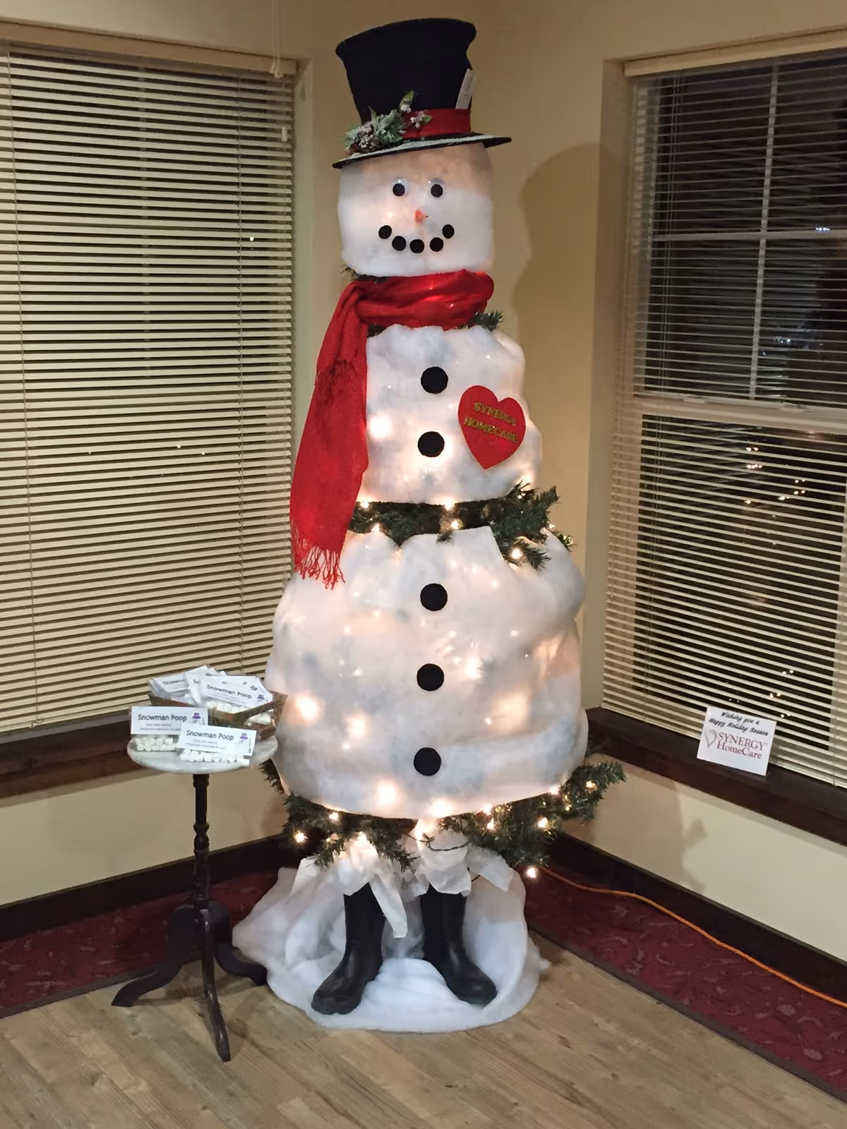 Tall illuminated snowman decoration indoors beside windows with blinds and a red scarf