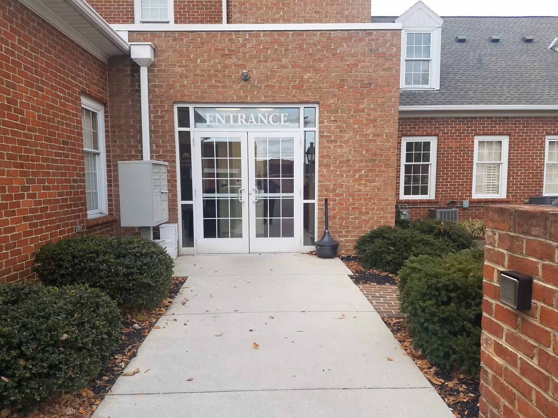 Brick building entrance with glass doors labeled 'ENTRANCE' and surrounding shrubs.