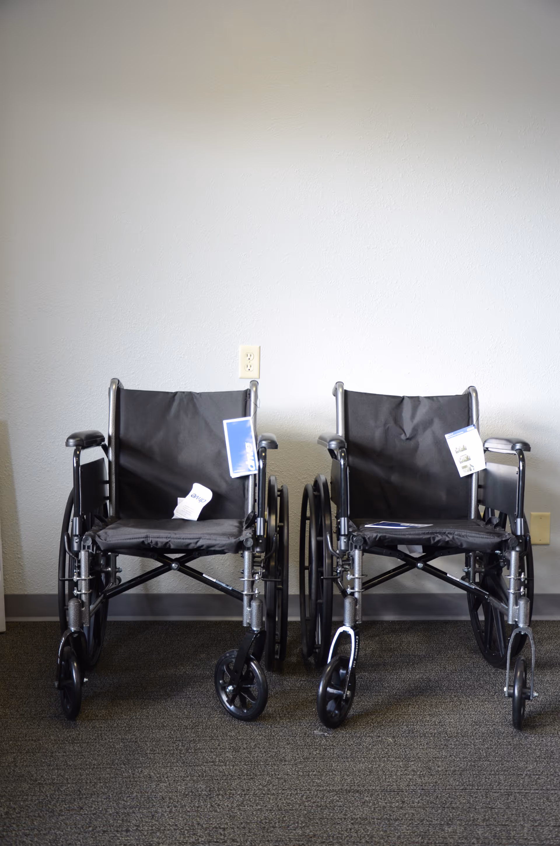Two black wheelchairs parked indoors against a plain wall.