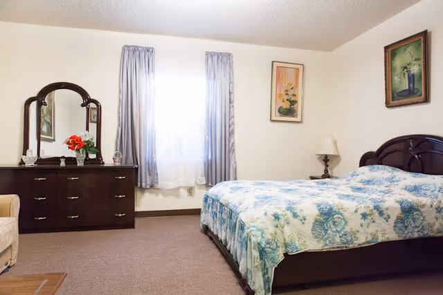 Traditional bedroom featuring a dark wood dresser with mirror, bed with floral quilt, and curtains.