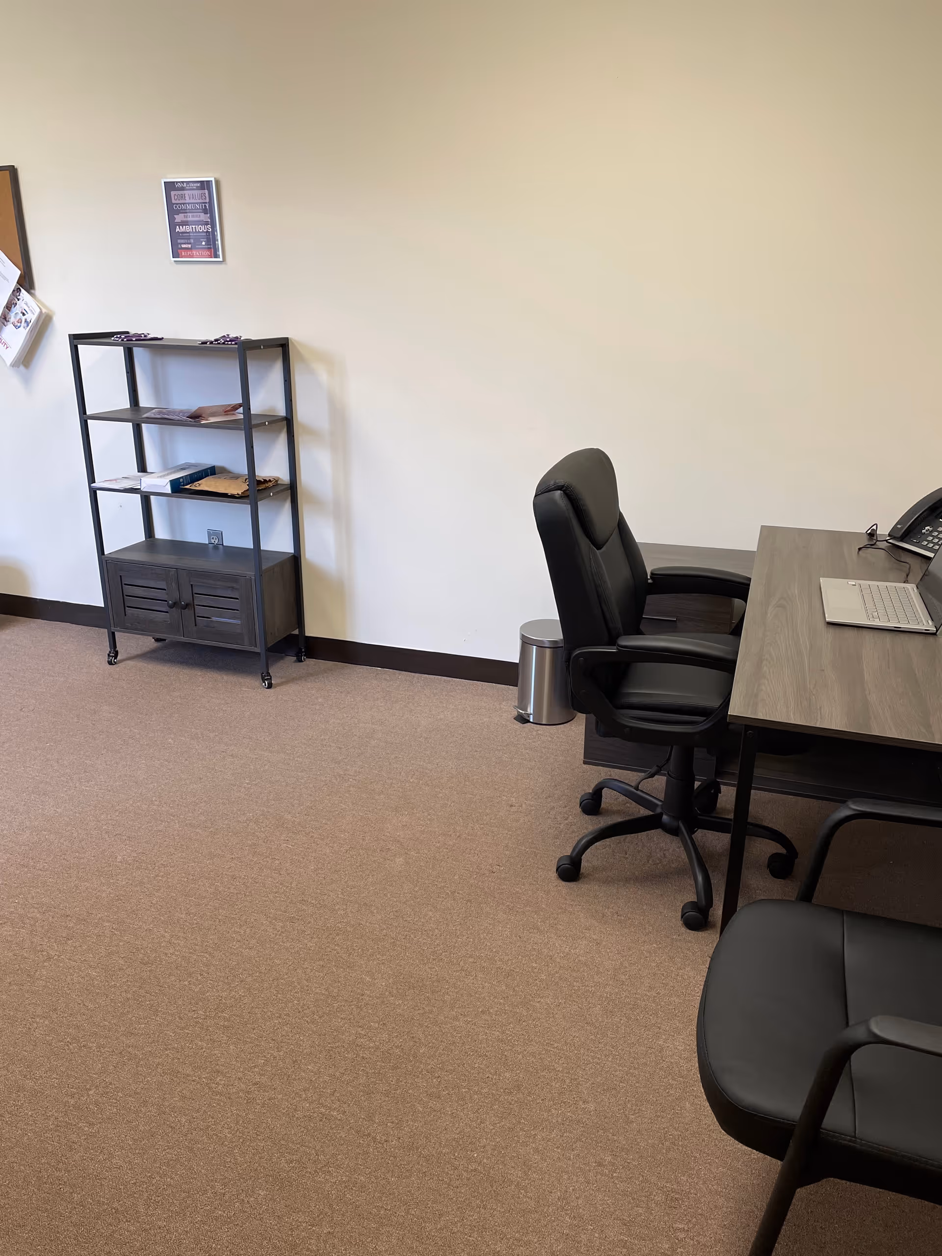 Office workspace with desk, chair, laptop, phone, and a metal shelving unit along wall.