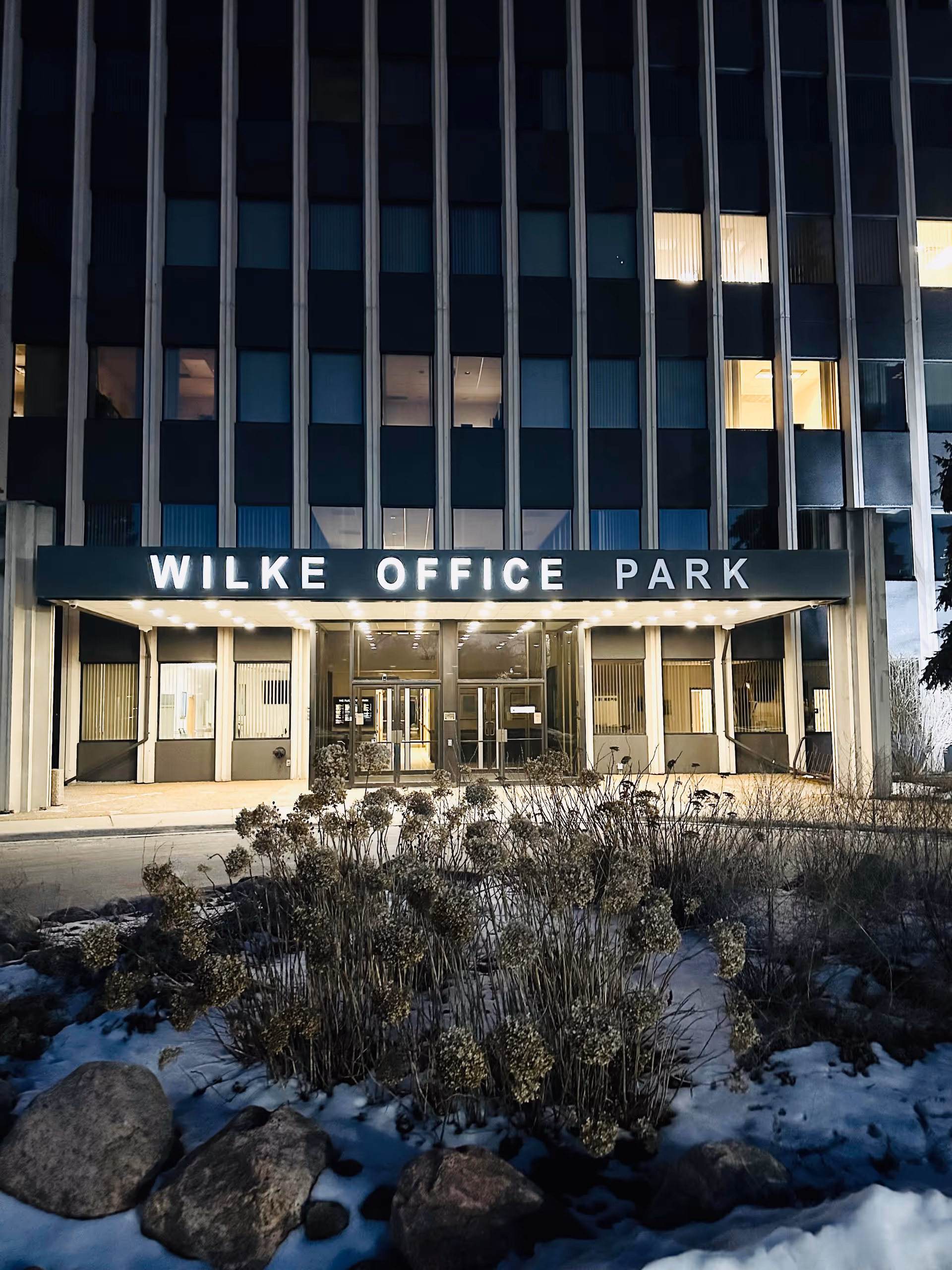 Night view of a building facade with a lit front entrance and Wilke Office Park sign.