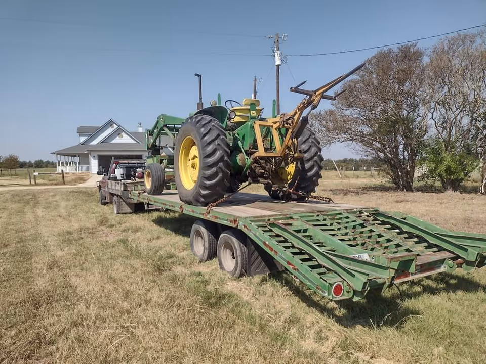 Flatbed trailer carrying a green tractor in a rural field near a white house.