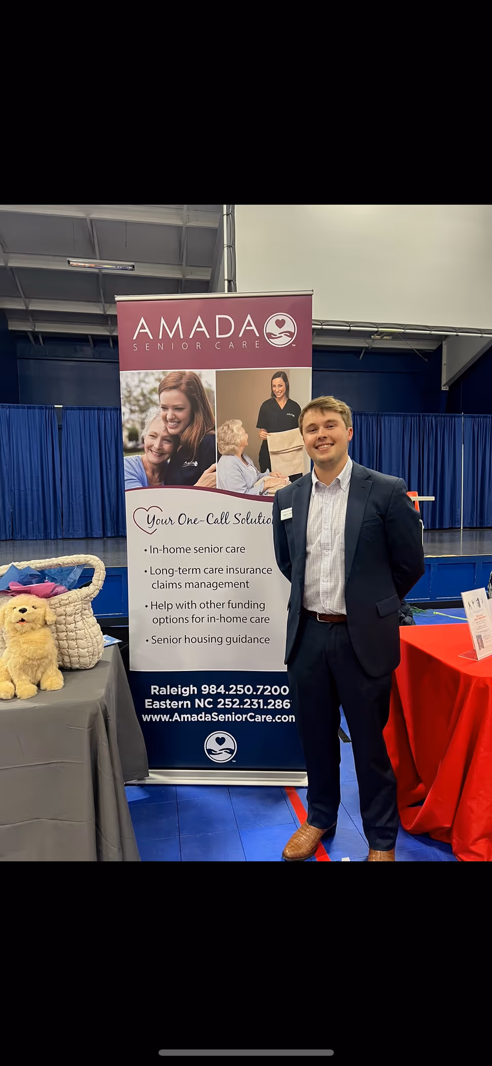 A man in a suit stands beside an Amada Senior Care banner at a booth.