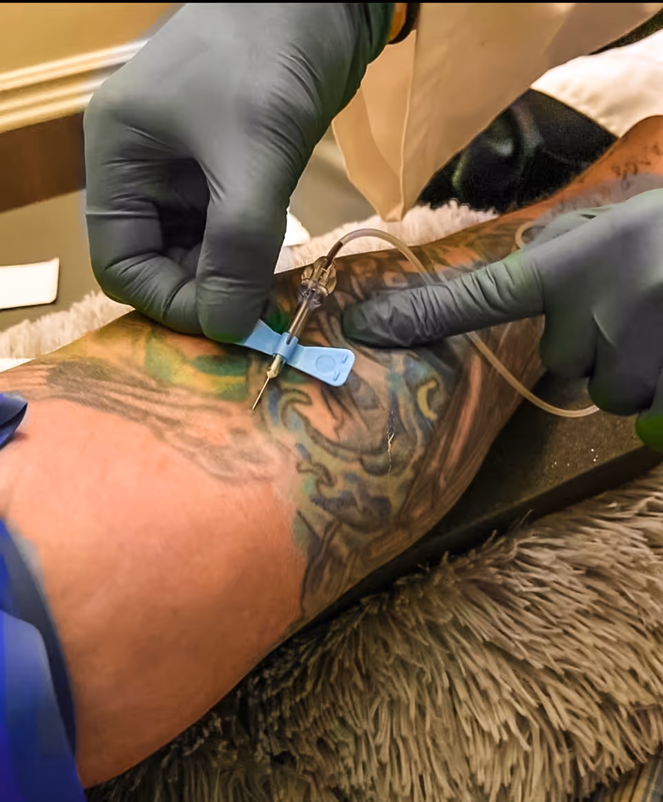 Gloved hands perform phlebotomy on a tattooed arm.