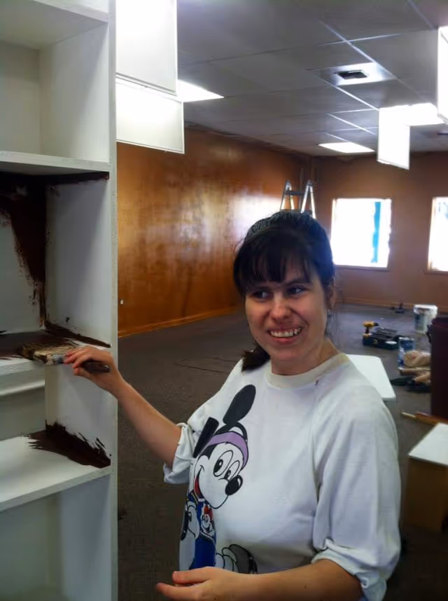 A woman applies brown paint to white shelves inside a large renovation room.