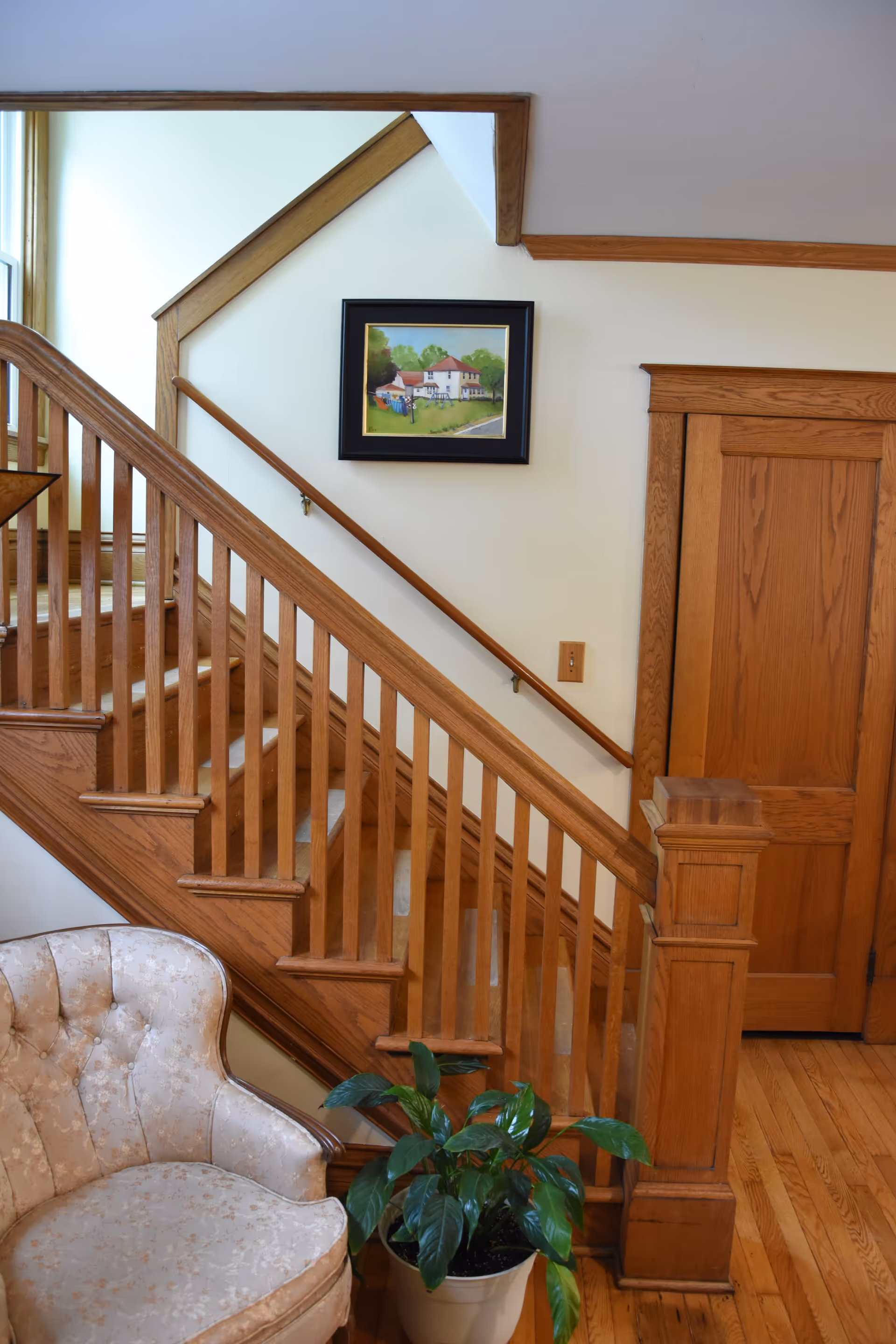 Wood staircase with railing, framed landscape painting, potted plant, and an upholstered chair in a foyer.