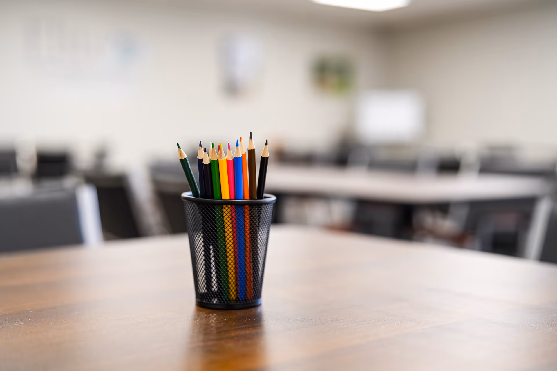 A black mesh pencil cup with colored pencils sits on a wooden table indoors.