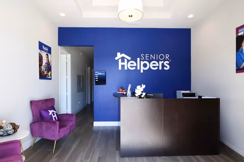 Reception area of Senior Helpers facility with blue wall logo, dark desk, and purple chairs.