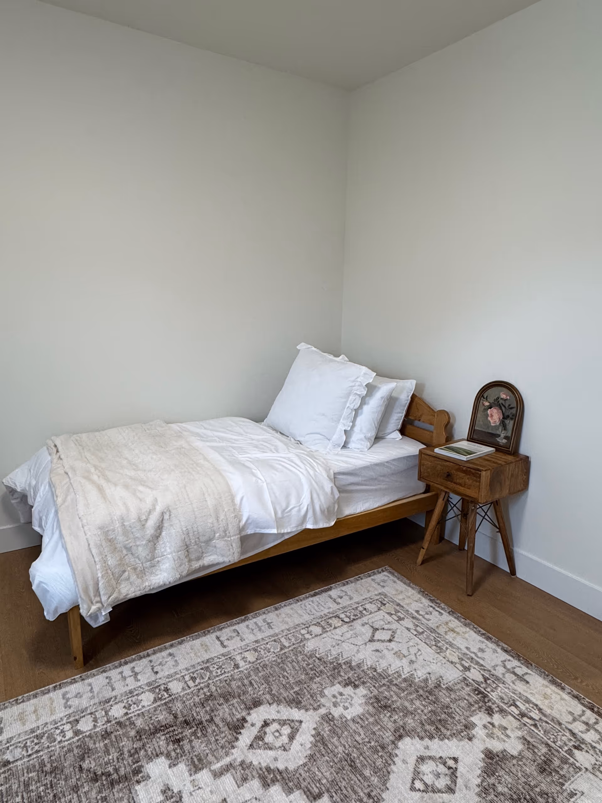 Single bed with white linens and a wooden nightstand in a simple bedroom.