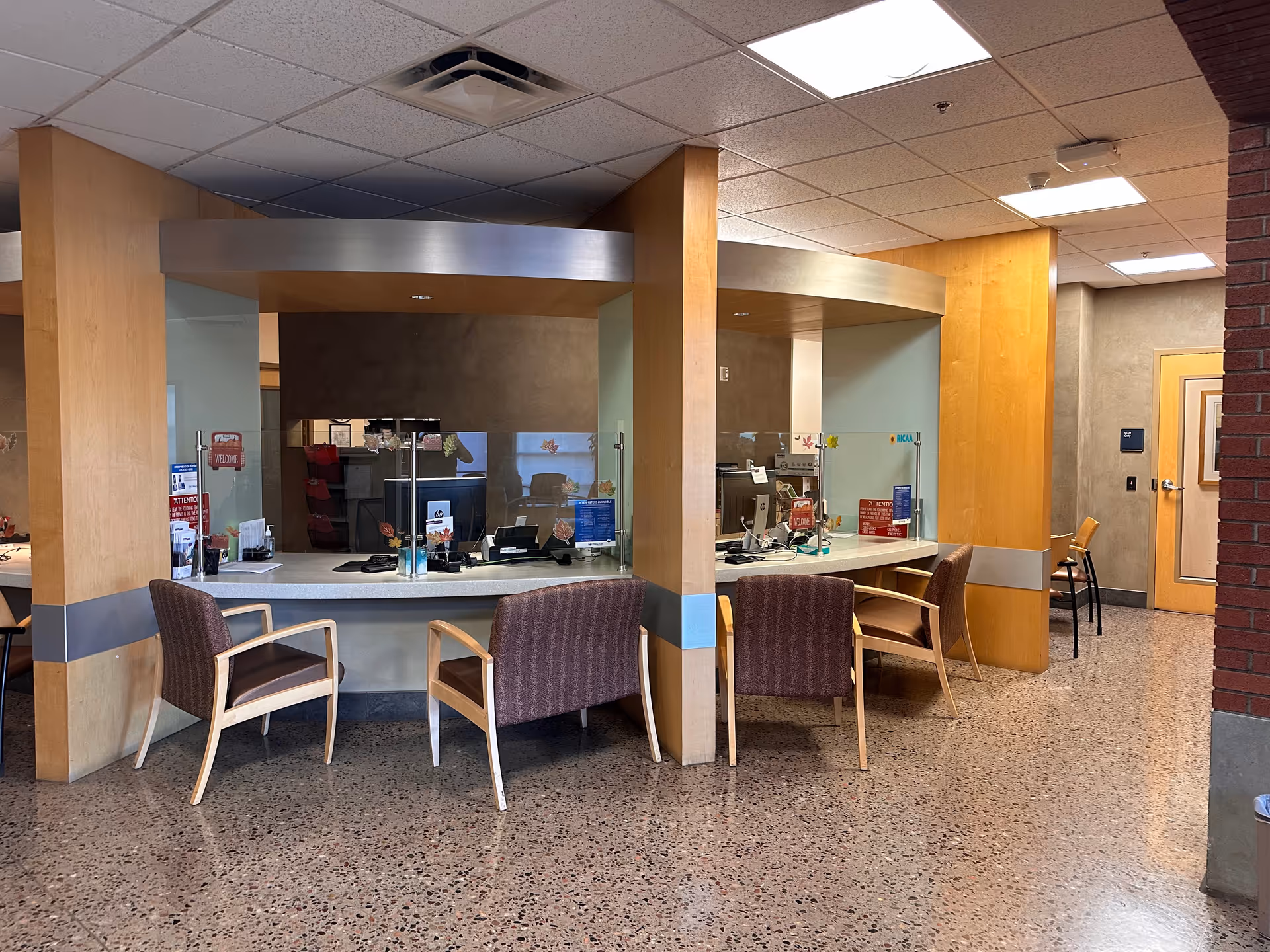 Interior hospital lobby with curved counters, glass barriers, and empty chairs.