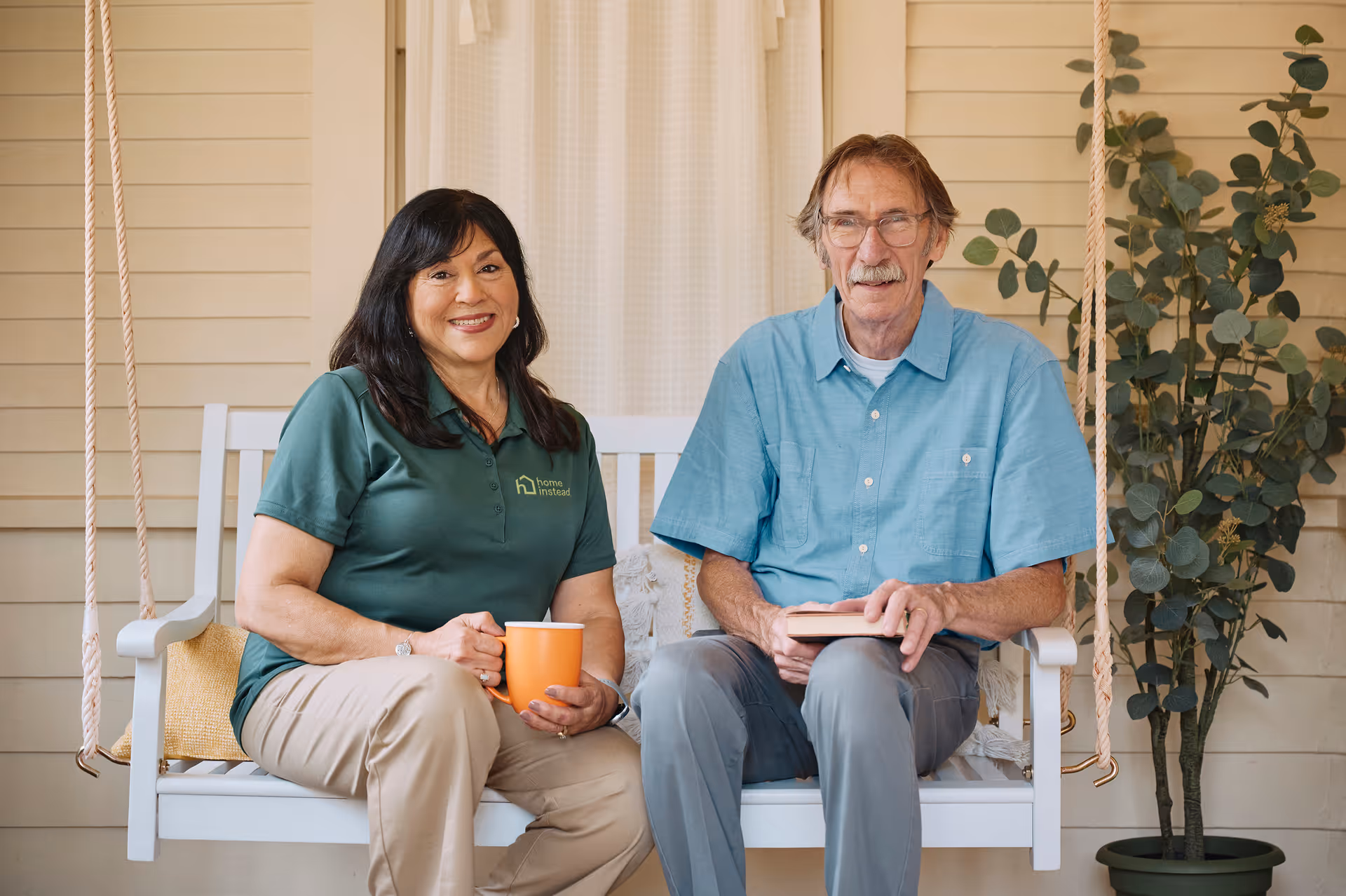 Two caregivers sit on a porch swing with an older man, smiling, mug and book.