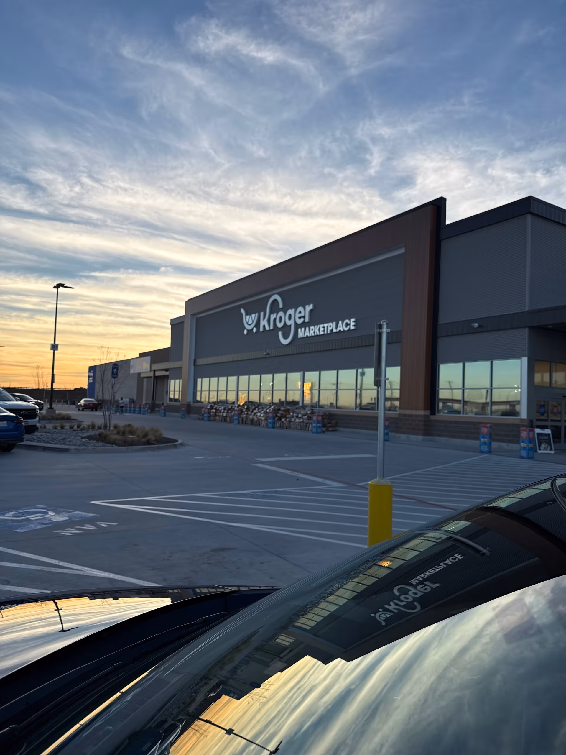 Exterior view of a Kroger Marketplace storefront at sunset, with parked cars.