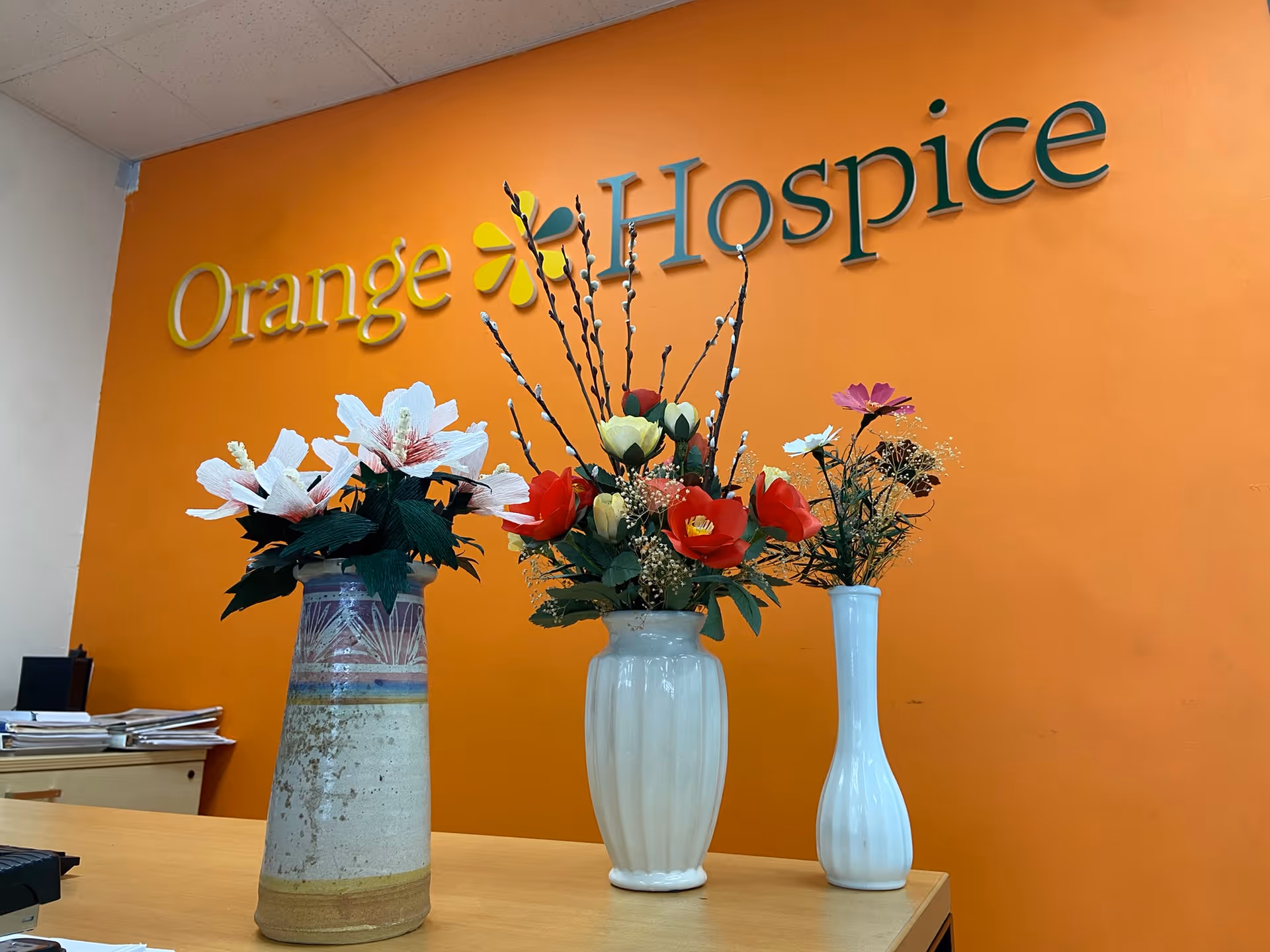Interior desk area with orange wall, Orange Hospice sign, and flower arrangements.
