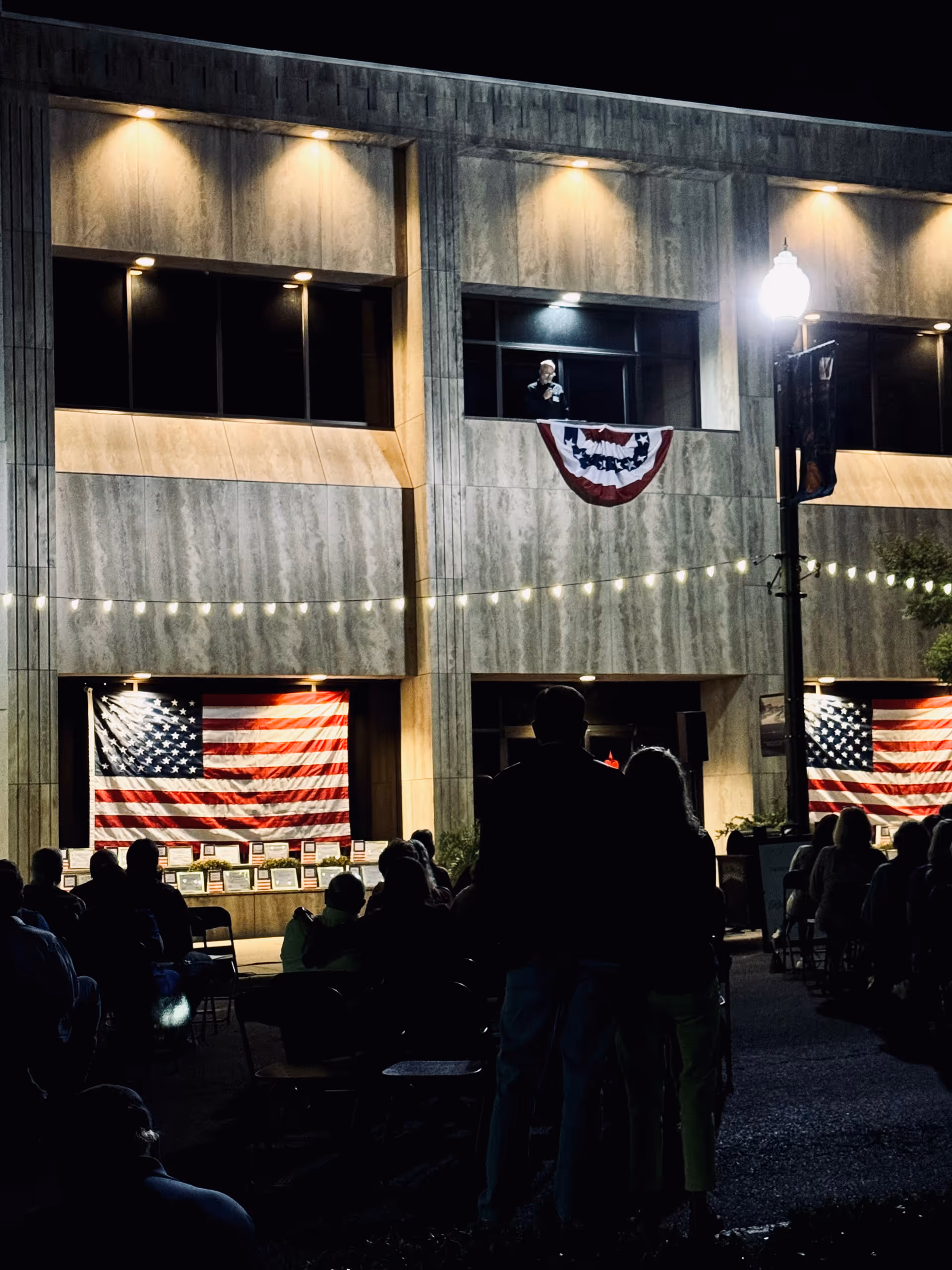 Night exterior of Heartlite Hospice Alabama building with American flags and attendees seated outside.