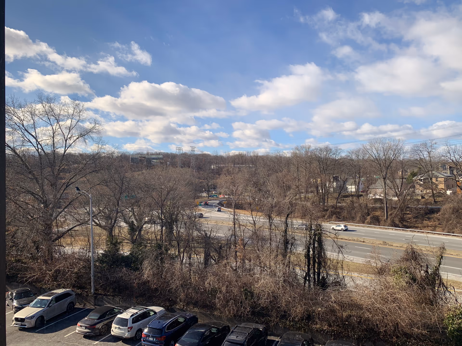 Parking lot with leafless trees, a highway, and blue sky beyond a building.