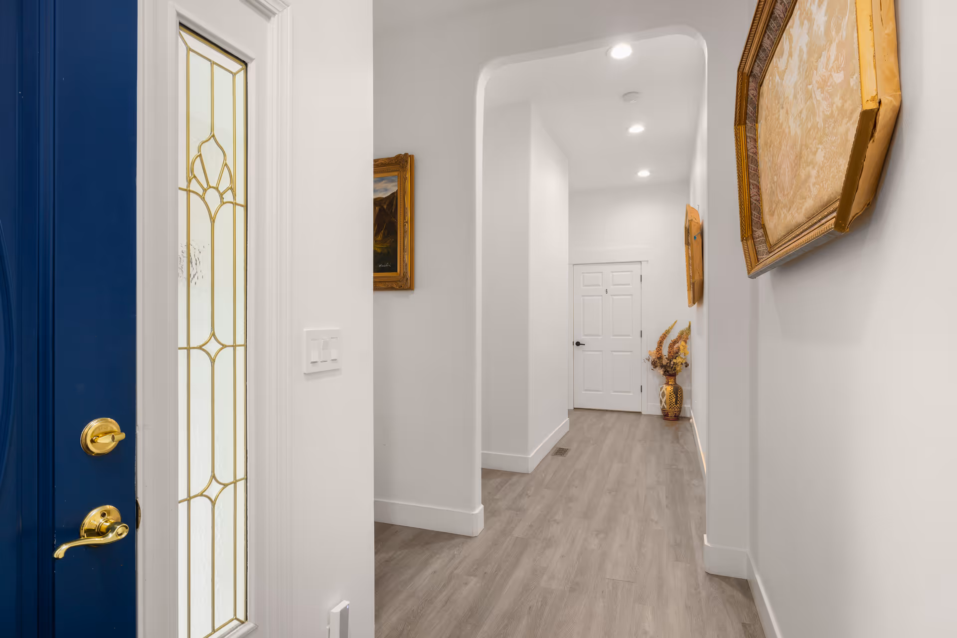 White hallway with arched doorway, blue front door, artwork, and wood-look flooring.
