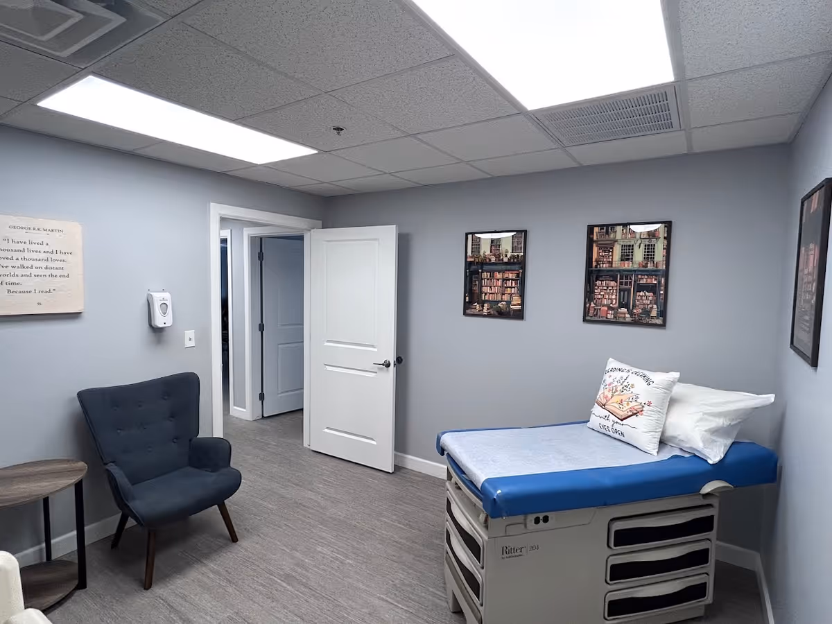 Medical examination room with blue padded table, chair, and wall art.